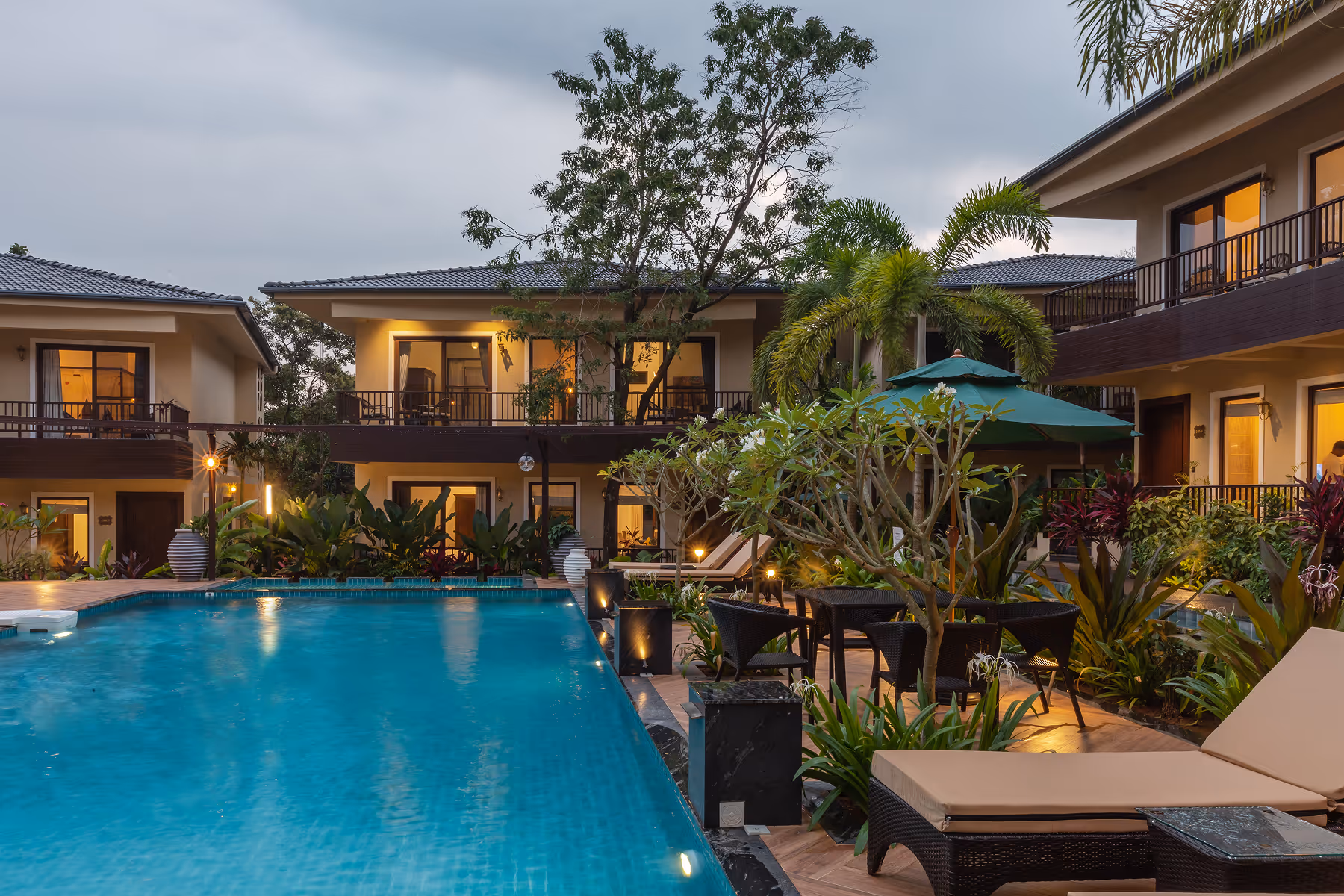 Outdoor swimming pool surrounded by lounge chairs, tables with umbrellas, tropical plants, and multi-story buildings with lit windows during dusk.
