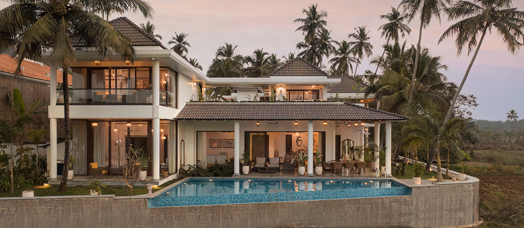 Modern two-story villa with tiled roof, outdoor seating, glass balcony, surrounded by palm trees, and a swimming pool in front.