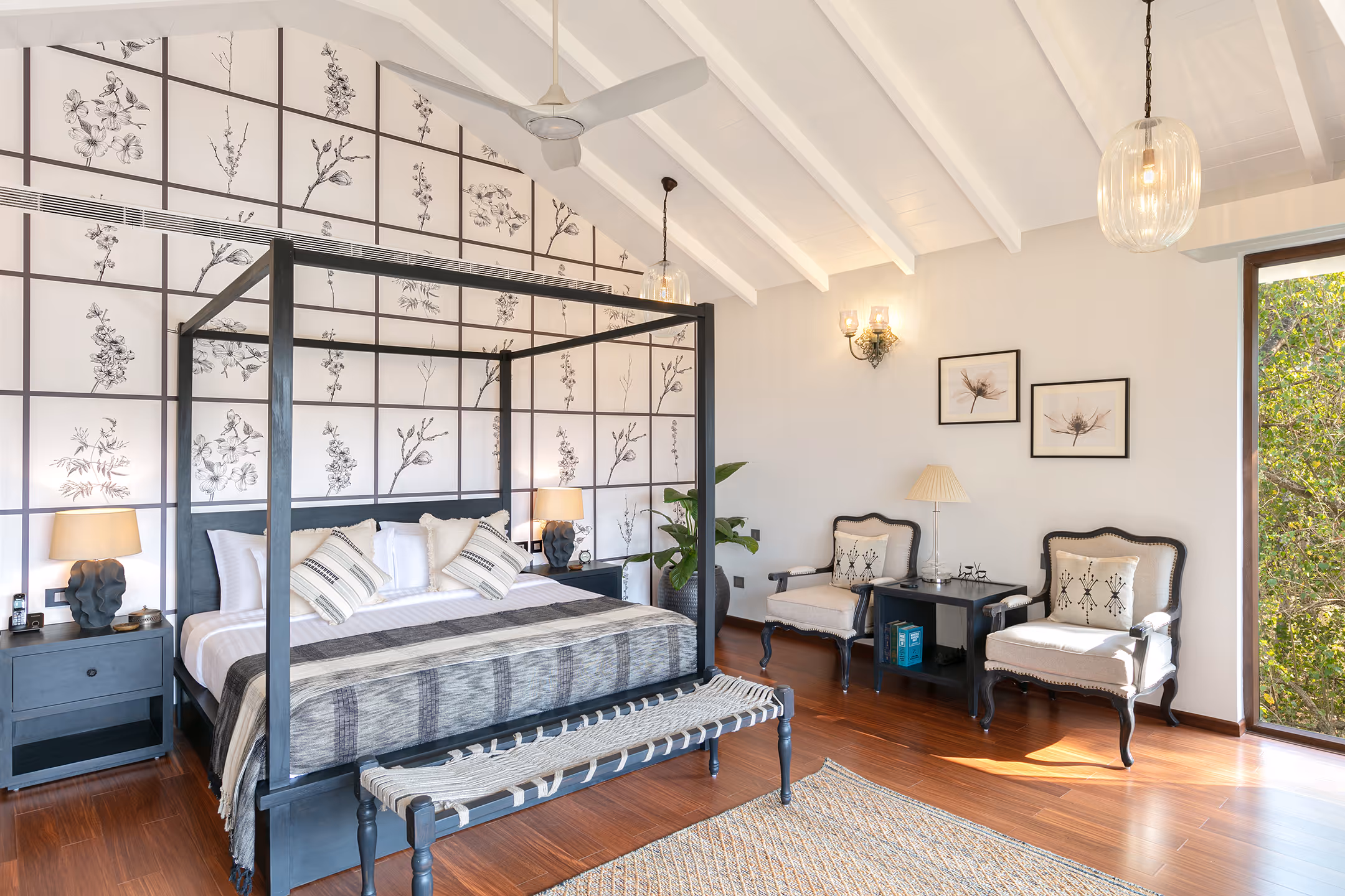 Bright bedroom with a black four-poster bed, floral wall decor, two armchairs, wooden floor, and large window showing greenery outside.