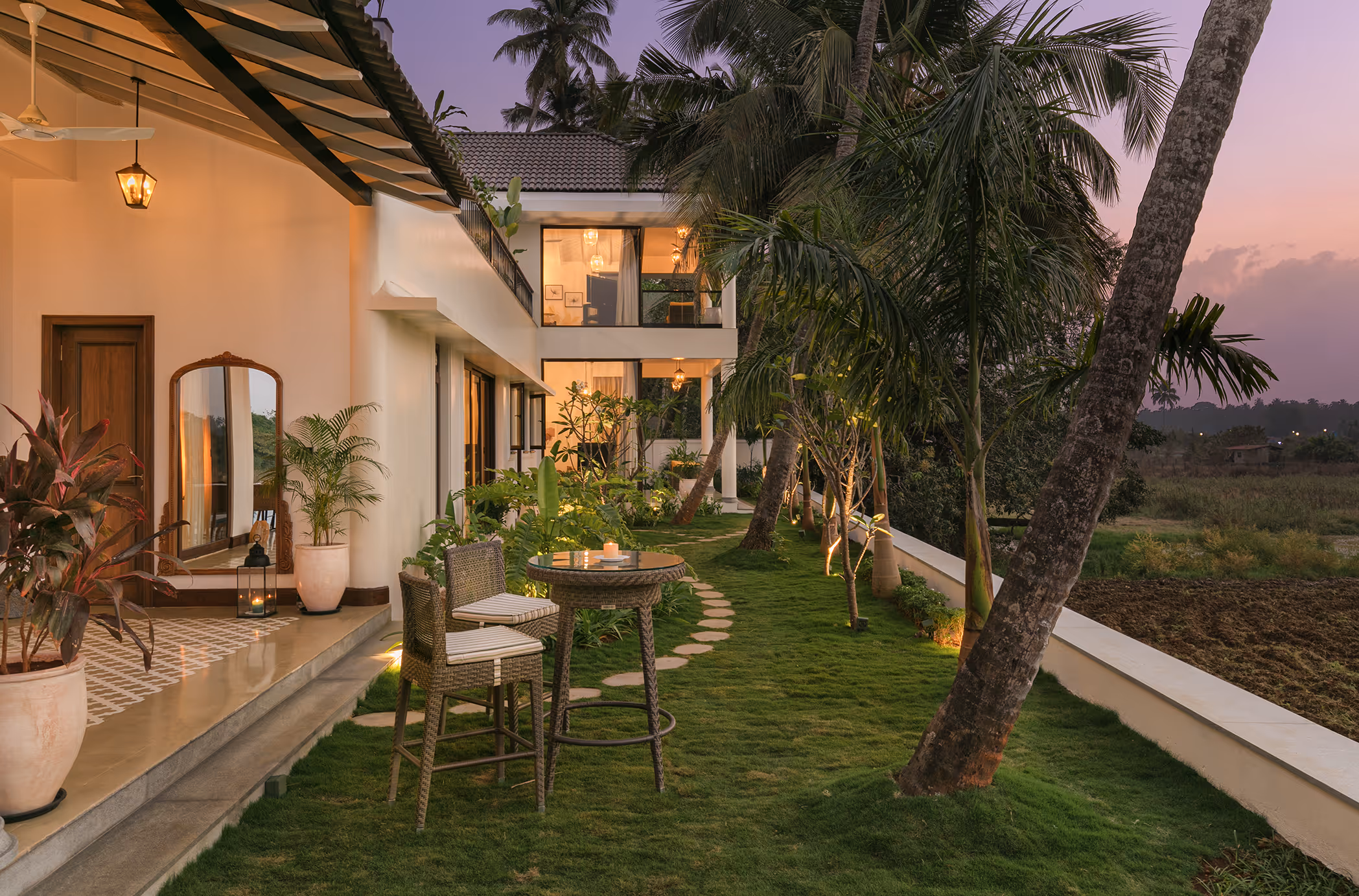 Outdoor patio area with wicker chairs and table on grassy lawn beside a modern white house, surrounded by palm trees at dusk.