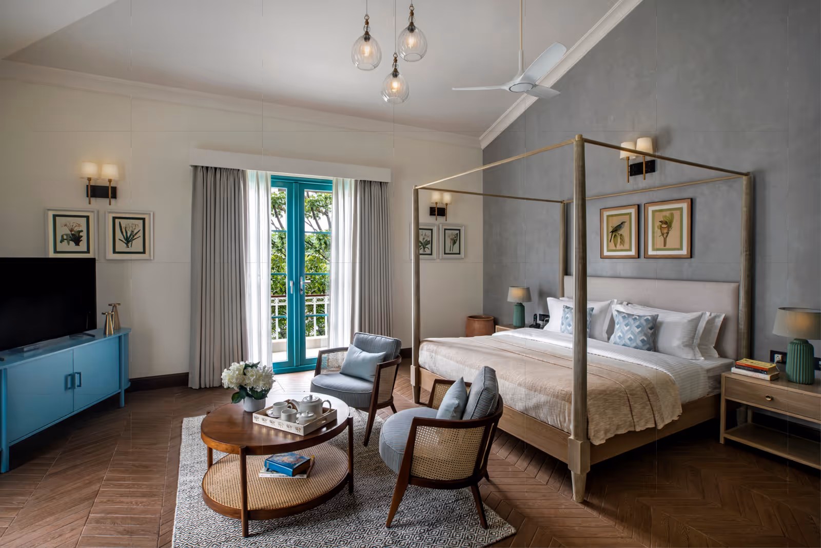 Modern bedroom with a four-poster bed, two gray upholstered chairs around a wooden coffee table, a blue media console with a TV, and turquoise-framed glass doors with curtains.