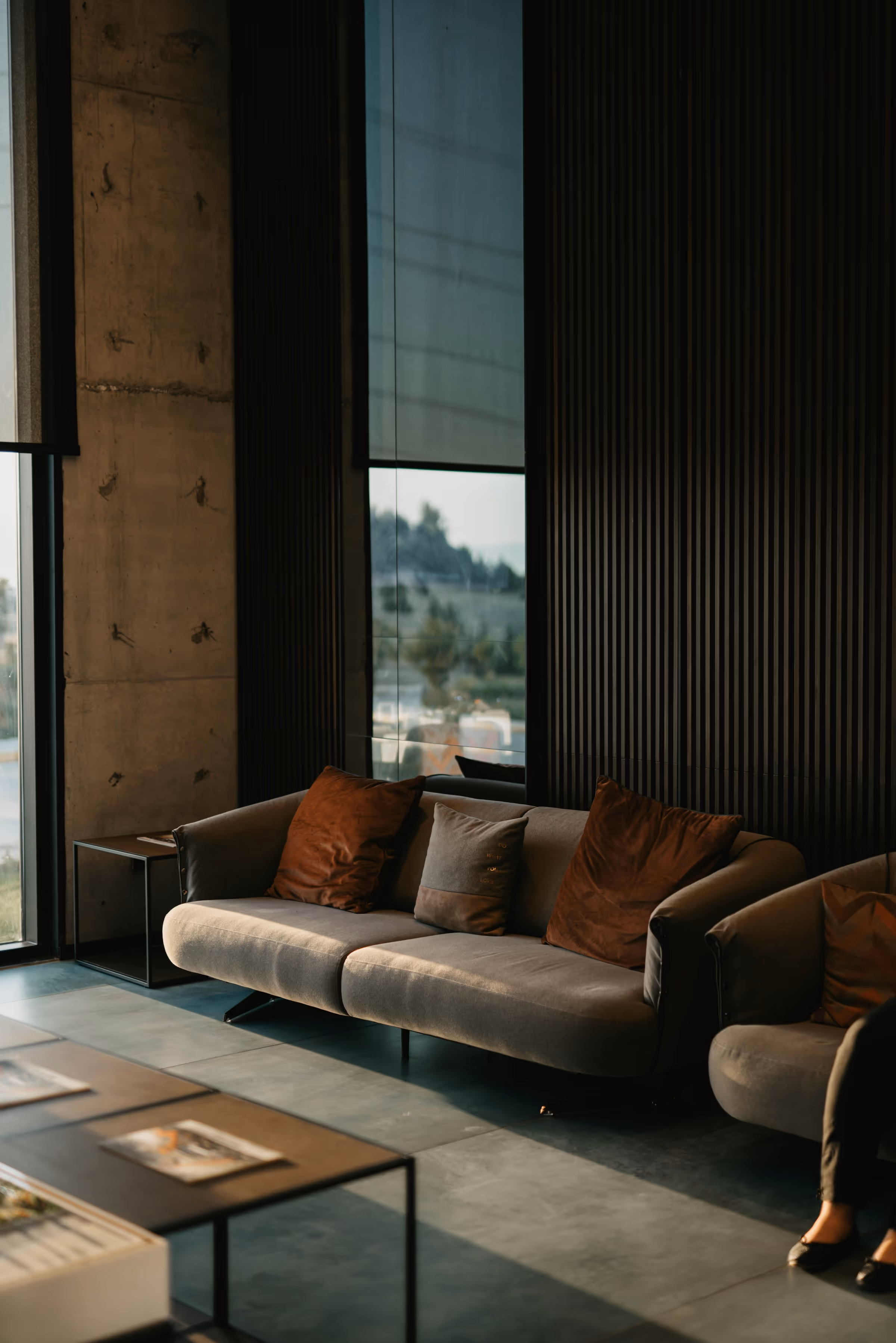 Modern living room with gray sofa and brown cushions beside large windows and black panel walls.