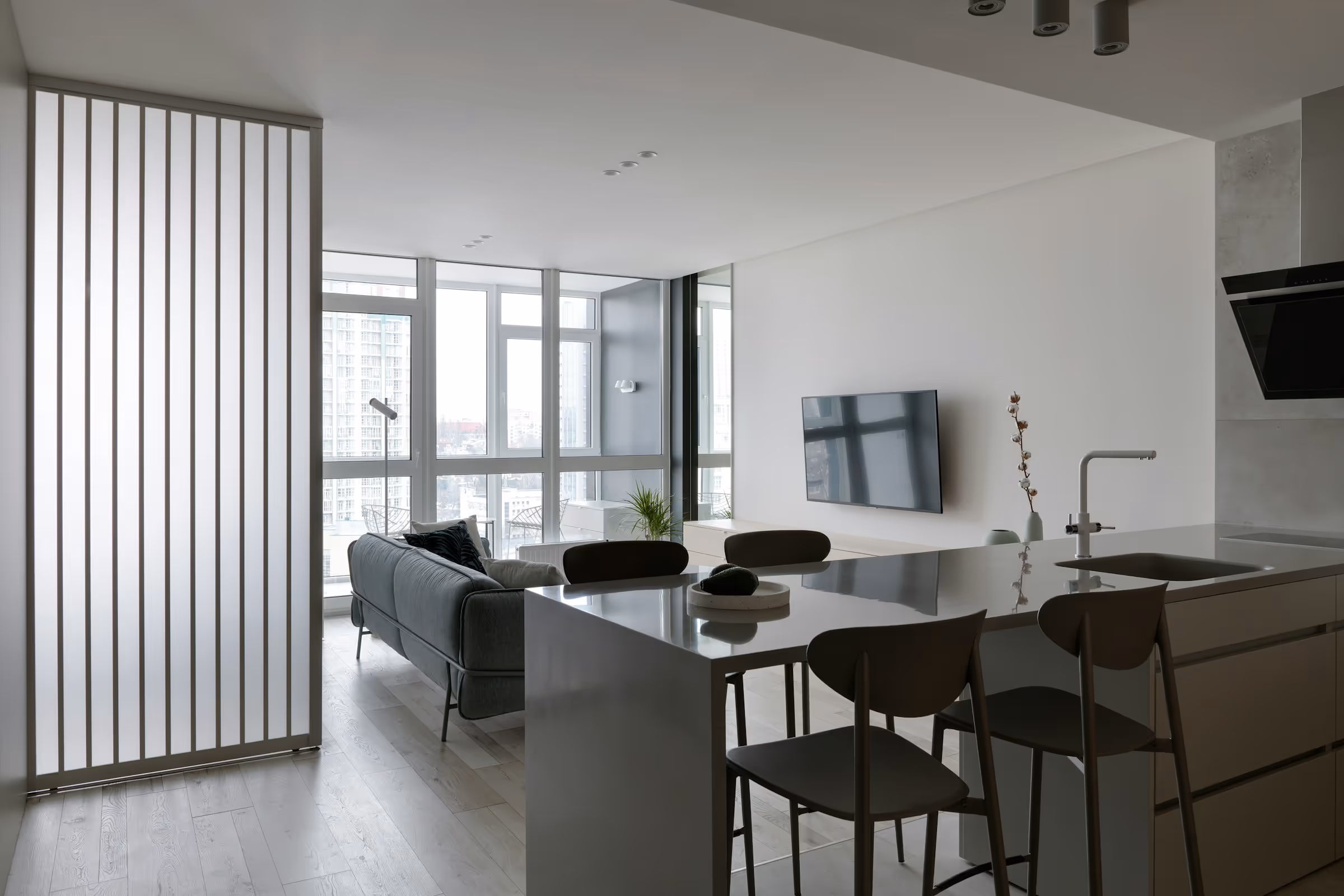 Modern open-plan living room and kitchen with large windows, a gray sofa, wall-mounted TV, and a kitchen island with chairs.