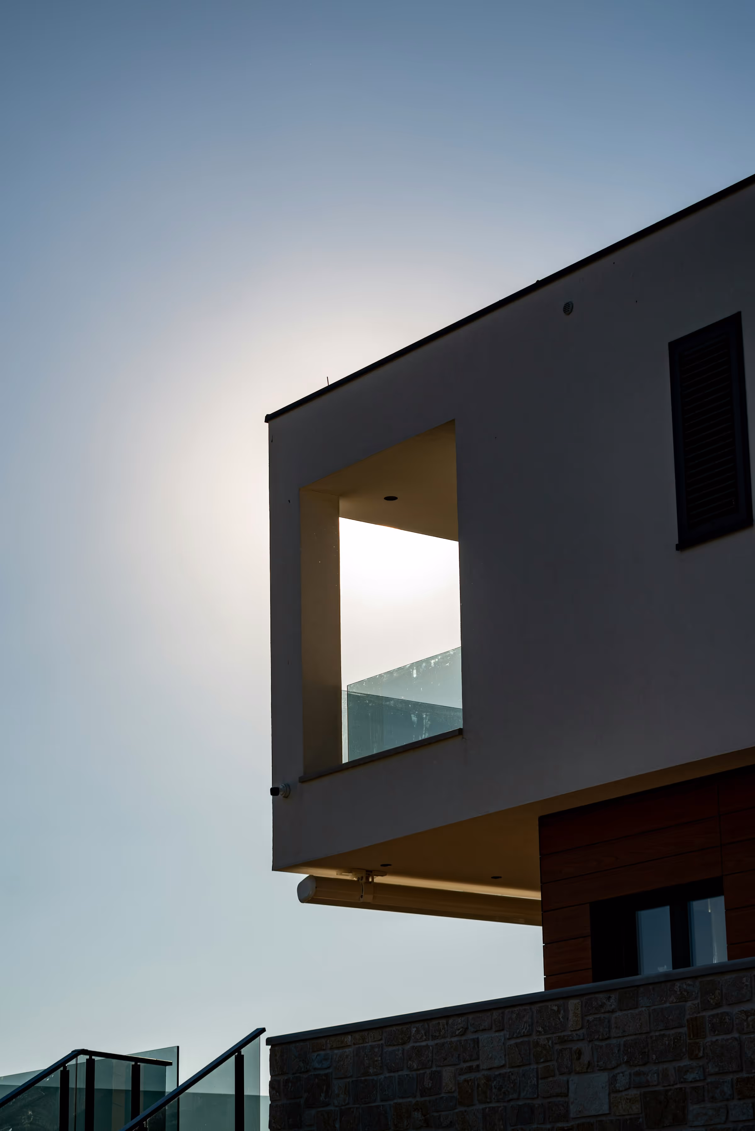 Modern building corner with a glass balcony and sunlight glowing behind it against a clear sky.