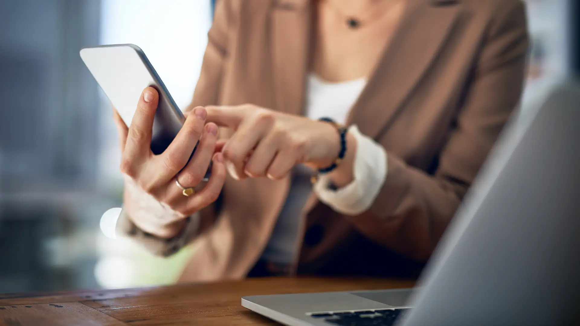 Person using a smartphone with a laptop on a wooden table nearby.