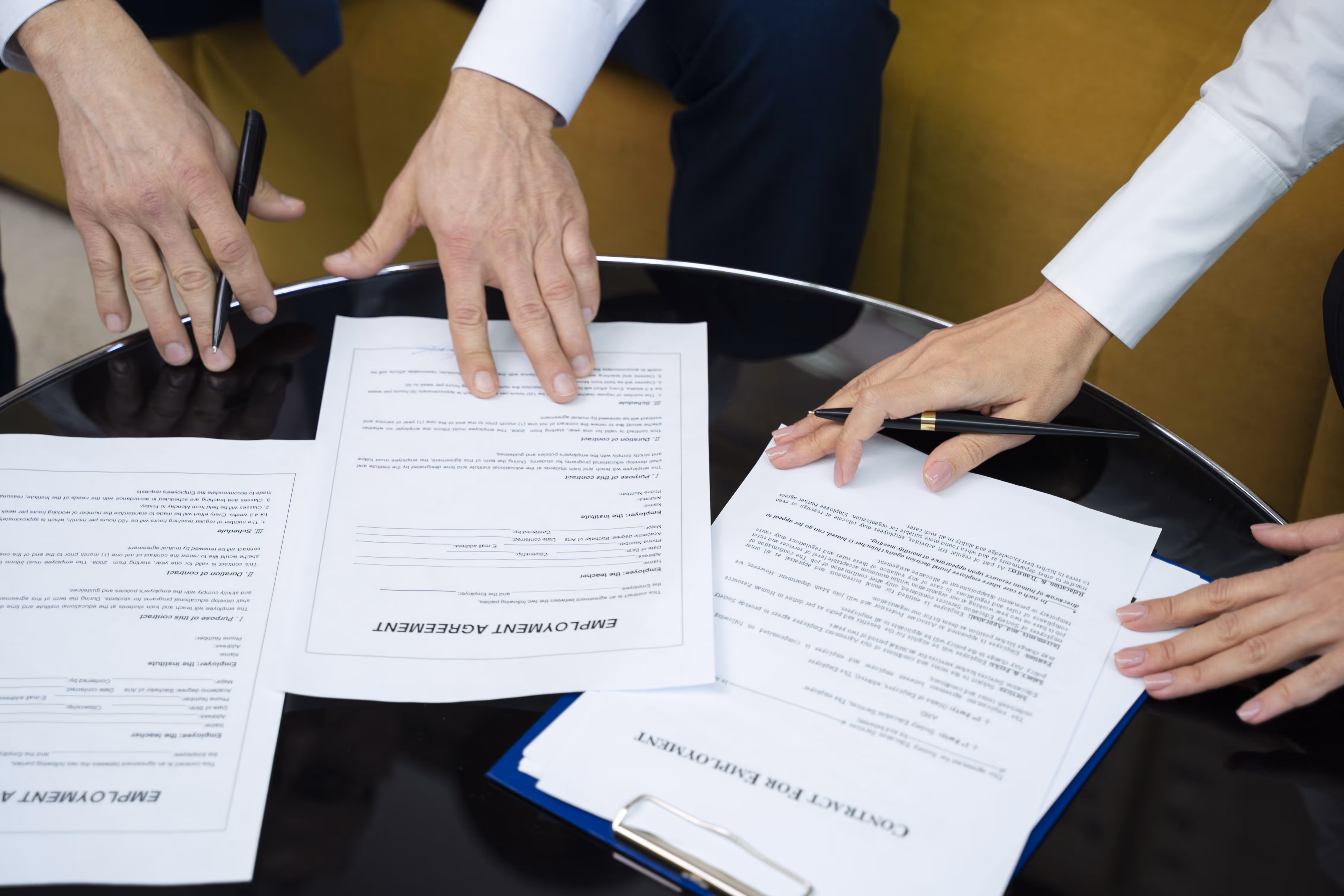 Hands signing a formal contract on a wooden desk with pen, paperwork, and clipboard.
