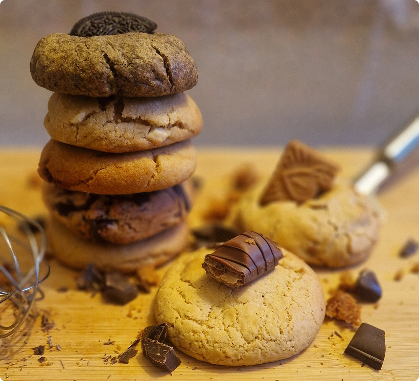Pile de cookies variés avec morceaux de chocolat et des biscuits sur une planche en bois.
