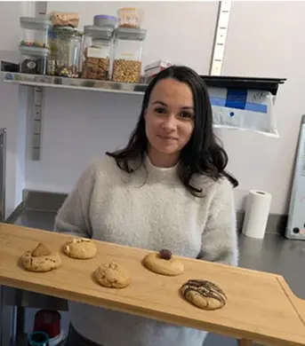 Femme souriante tenant une planche en bois avec cinq biscuits variés sur une table de cuisine.