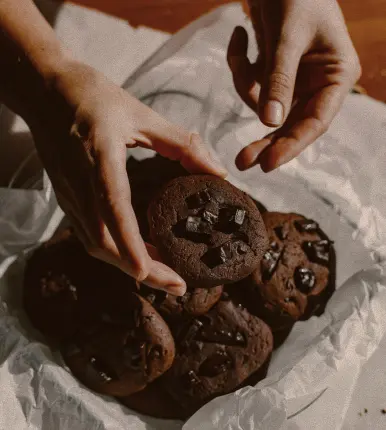 Deux mains tiennent un cookie au chocolat foncé avec des morceaux de chocolat, au-dessus d'un contenant tapissé de papier blanc rempli de cookies similaires.