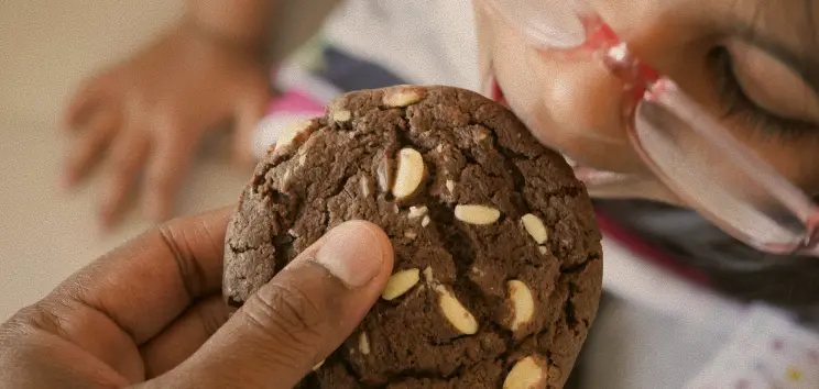 Une main tenant un biscuit au chocolat avec des morceaux de noix, proche du visage d'un enfant avec des lunettes roses.