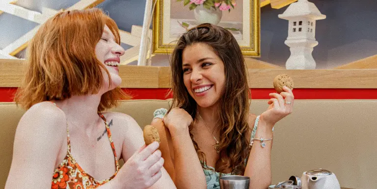 Deux femmes souriantes tenant des biscuits tout en discutant dans un café.