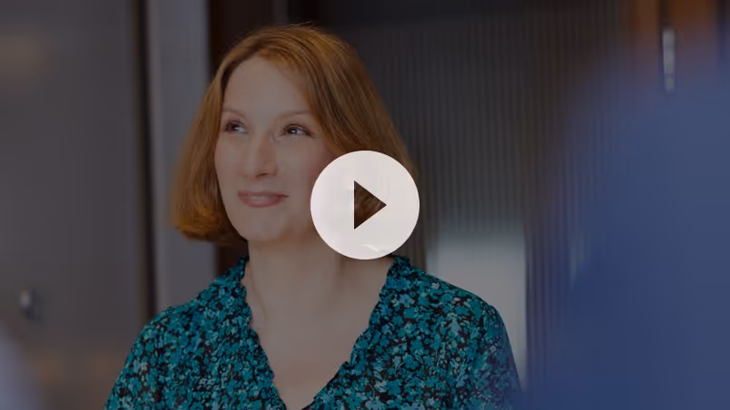 Smiling woman with short light brown hair wearing a teal floral blouse in an indoor setting.