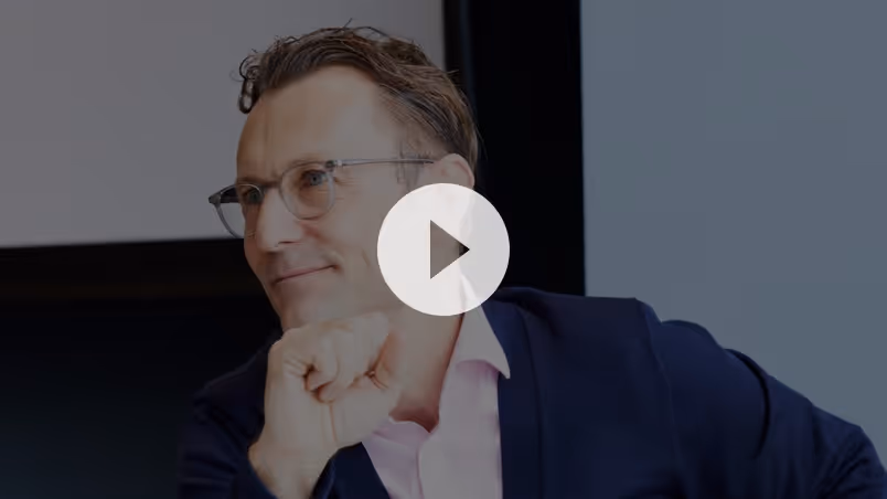 Man with glasses wearing a navy blazer and light pink shirt, resting his chin on his hand and smiling.
