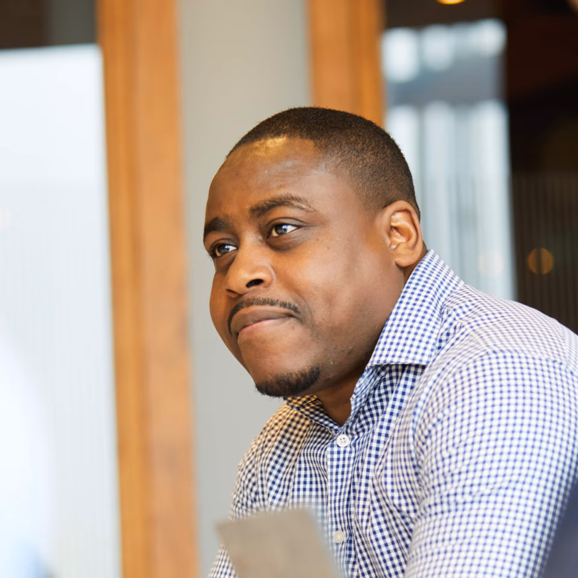 Man with short hair wearing a blue and white checkered shirt, looking thoughtfully to the side indoors.