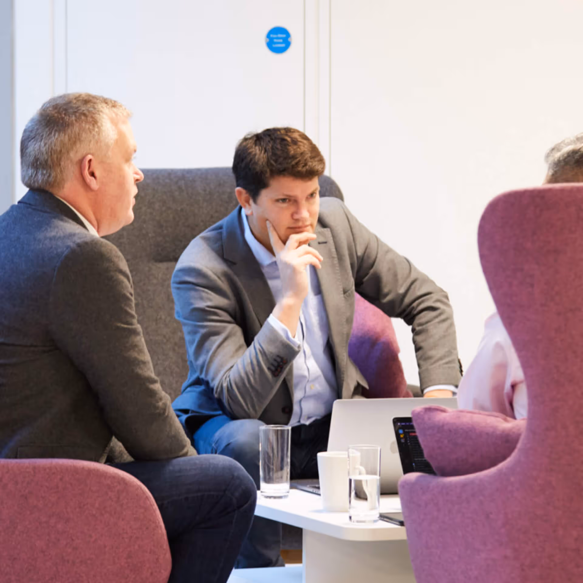 Three men in business attire having a focused discussion in a meeting room with laptops and glasses of water on a white table.