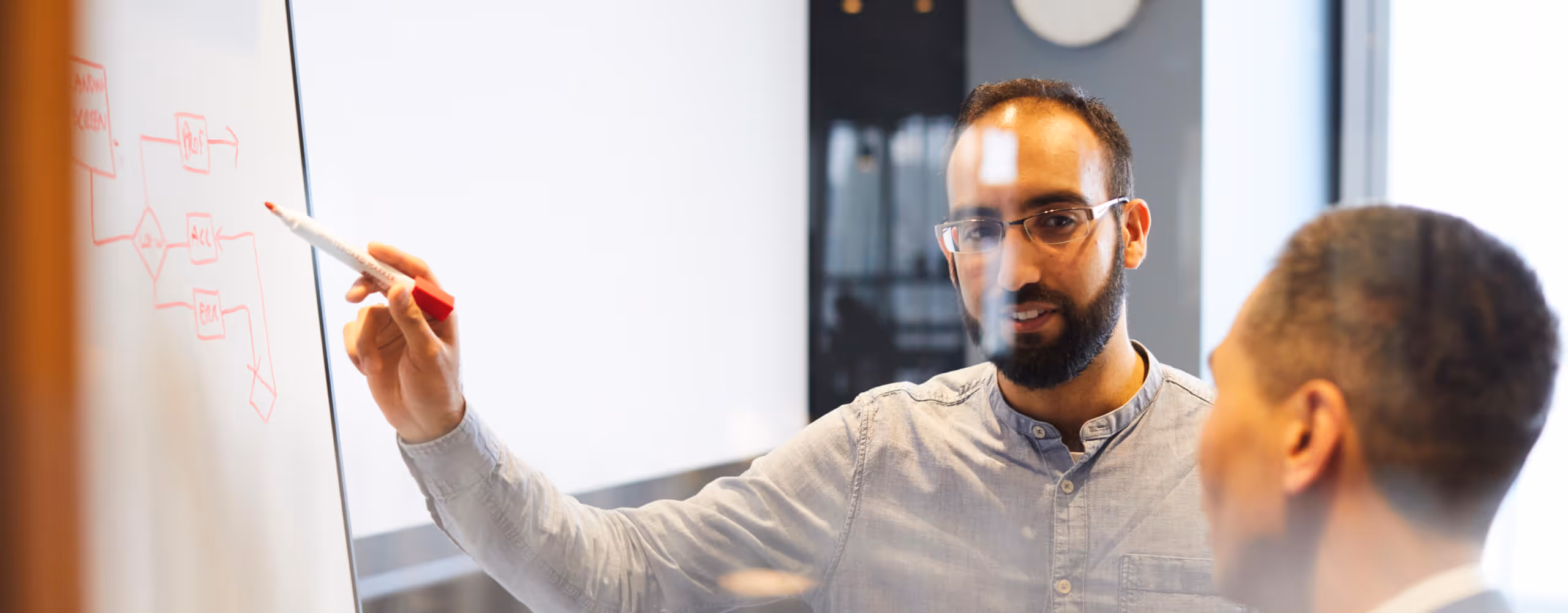 Man with glasses explaining a diagram on a whiteboard to another person in an office.