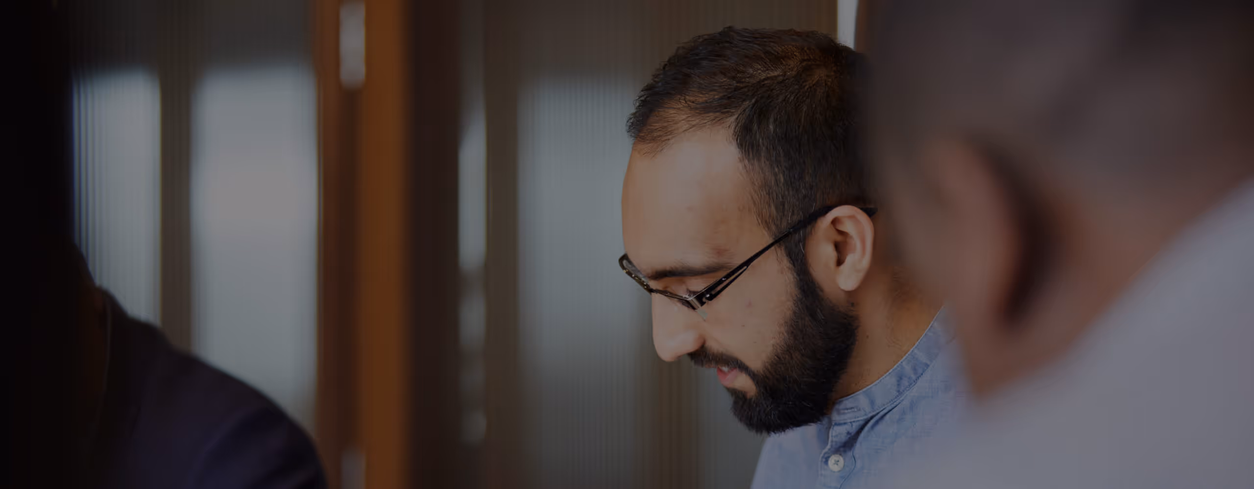 Man with glasses and a beard focusing on work or conversation in an indoor setting.