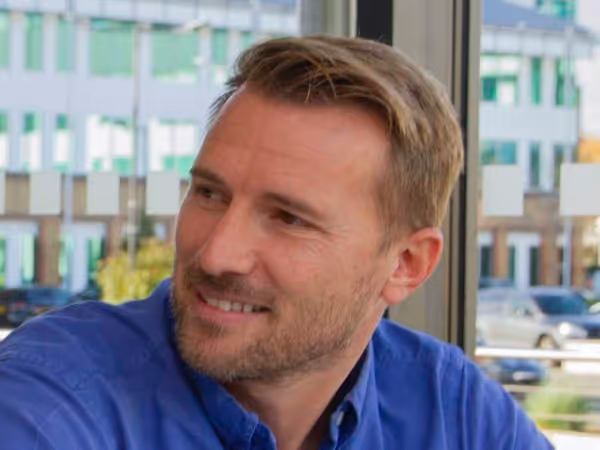 Smiling man with short light brown hair and beard, wearing a blue button-up shirt, sitting indoors near a window.