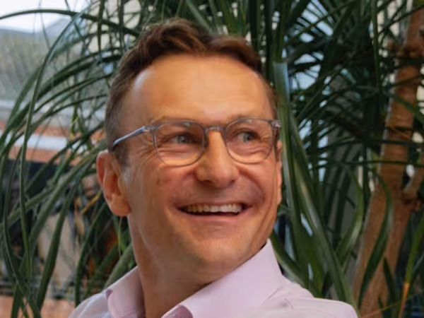 Smiling man wearing glasses and a light purple shirt with green plants in the background.