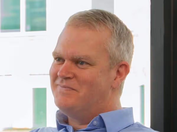 Smiling middle-aged man with short gray hair wearing a light blue collared shirt, sitting indoors near a window.