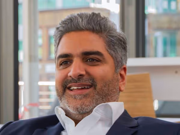 Smiling man with salt-and-pepper hair and beard wearing a white shirt and dark suit jacket indoors.