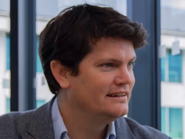 Man with brown hair wearing a gray blazer and blue shirt looking to the right in an office setting.