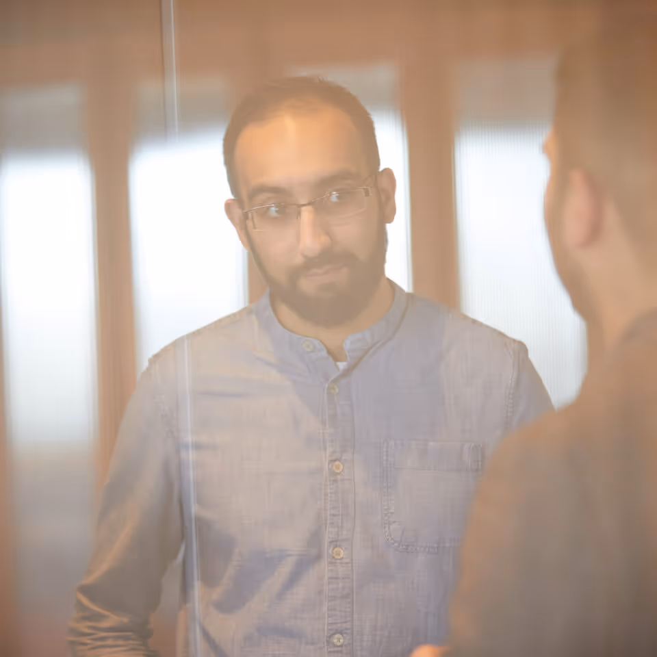 Man with glasses and beard wearing a light blue shirt looking at his reflection in a mirror.