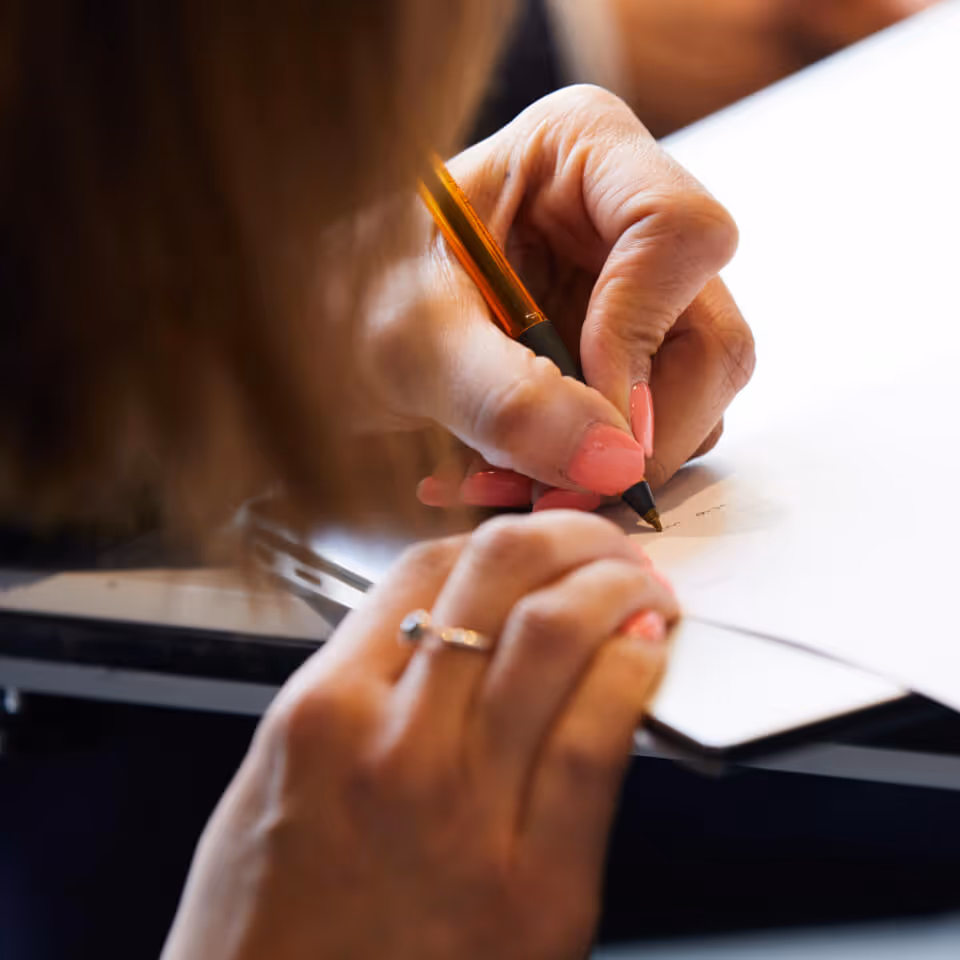 Close-up of a person with pink-painted nails writing on paper with a pen, holding the paper with the other hand wearing a ring.