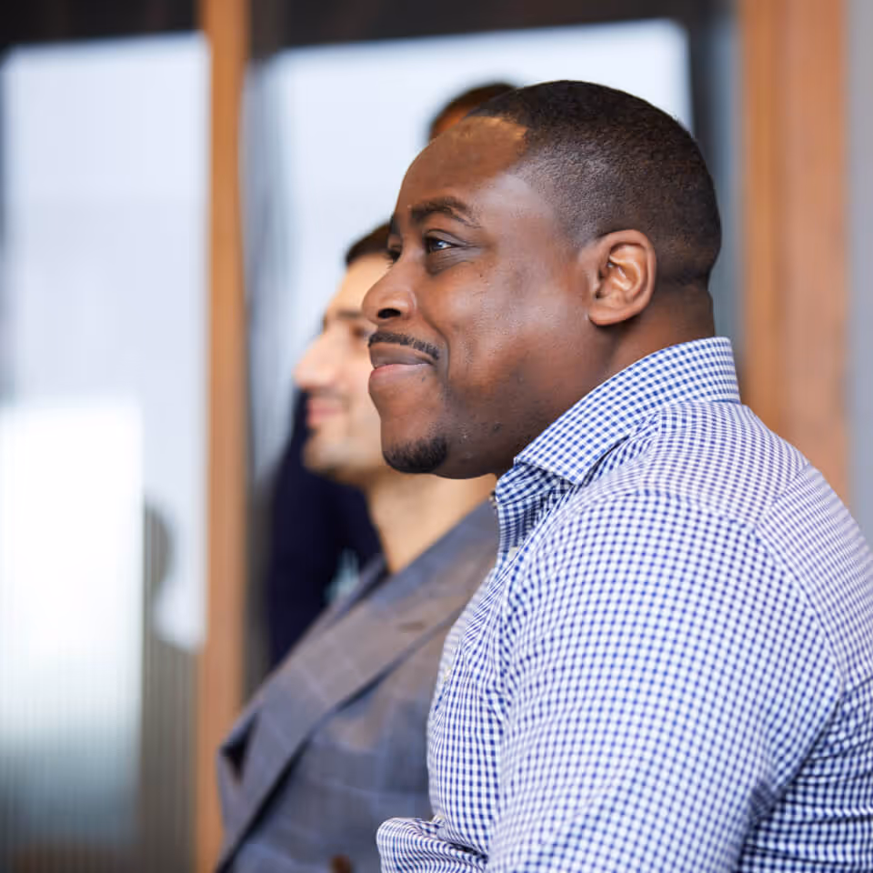 Three men attentively listening in a professional setting, focusing on the man in front wearing a checked shirt.