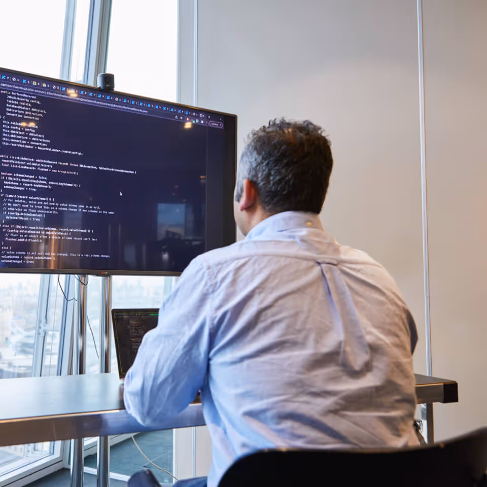 Person with dark hair in a light blue shirt sitting at a desk and looking at a large monitor displaying computer code in an office with large windows.