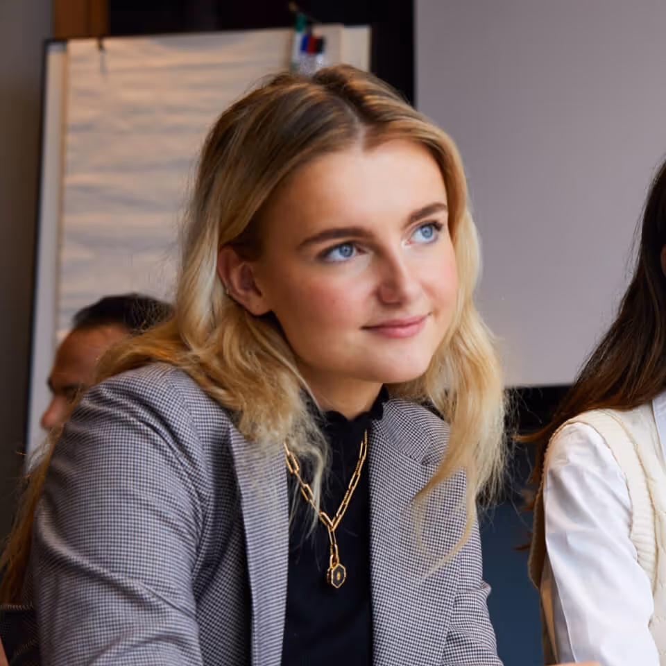 Young blonde woman with blue eyes wearing a checkered blazer and gold necklace in a meeting room.