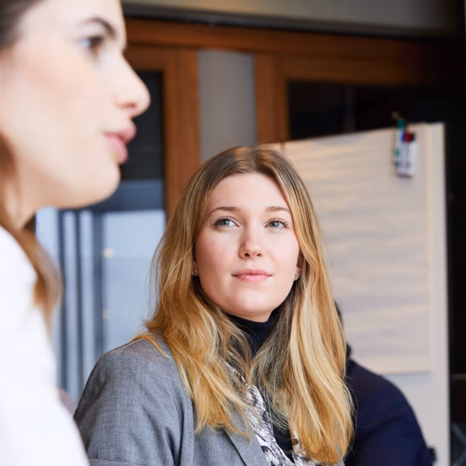 Blonde woman in a gray blazer attentively listening to another person speaking in an office setting.