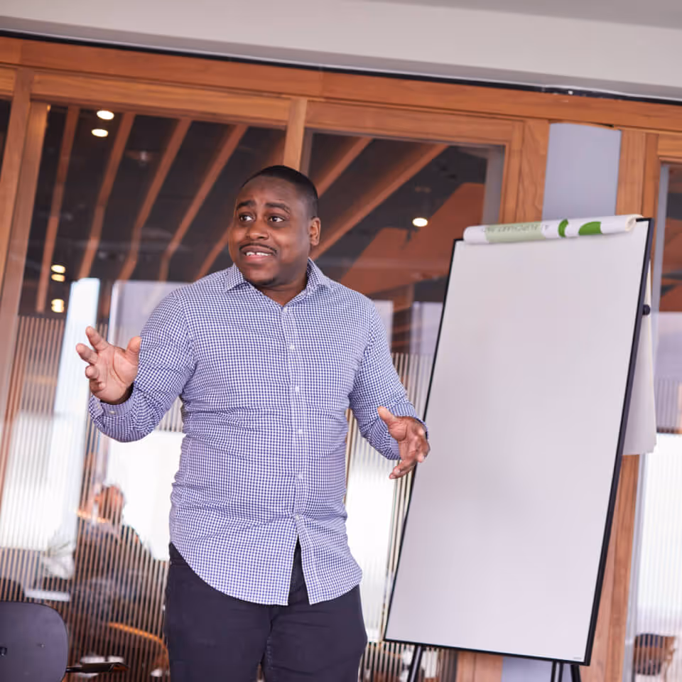 Man in checkered shirt gesturing as he speaks next to a blank flip chart in a modern office.