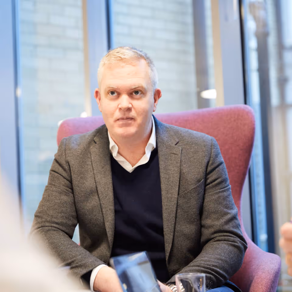 Man with short gray hair wearing a gray blazer and dark sweater sitting in a purple chair indoors.