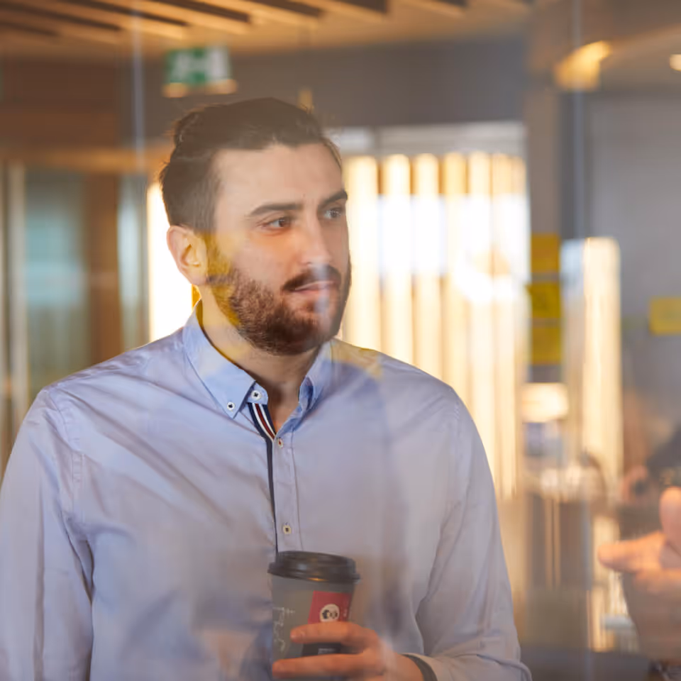 Man with dark hair and beard holding a coffee cup inside an office with blurred background.