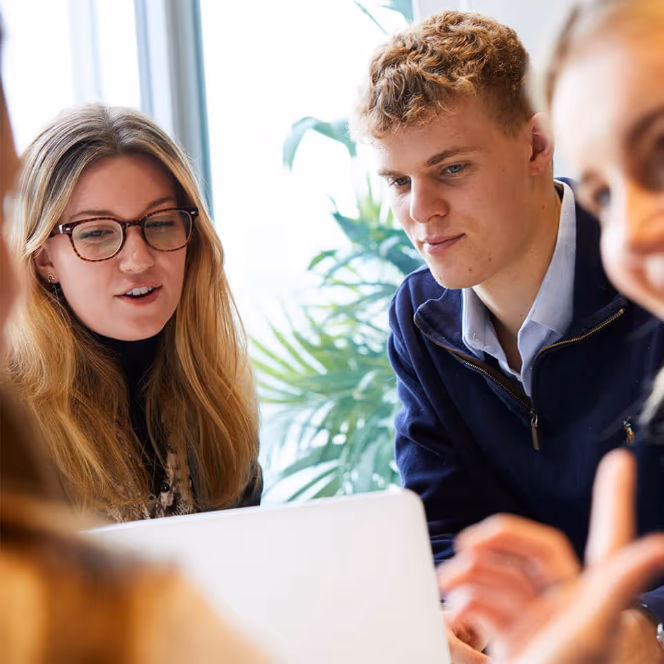 Two young adults looking at a laptop screen, engaged in a discussion, with a blurred person in the foreground and indoor plants in the background.
