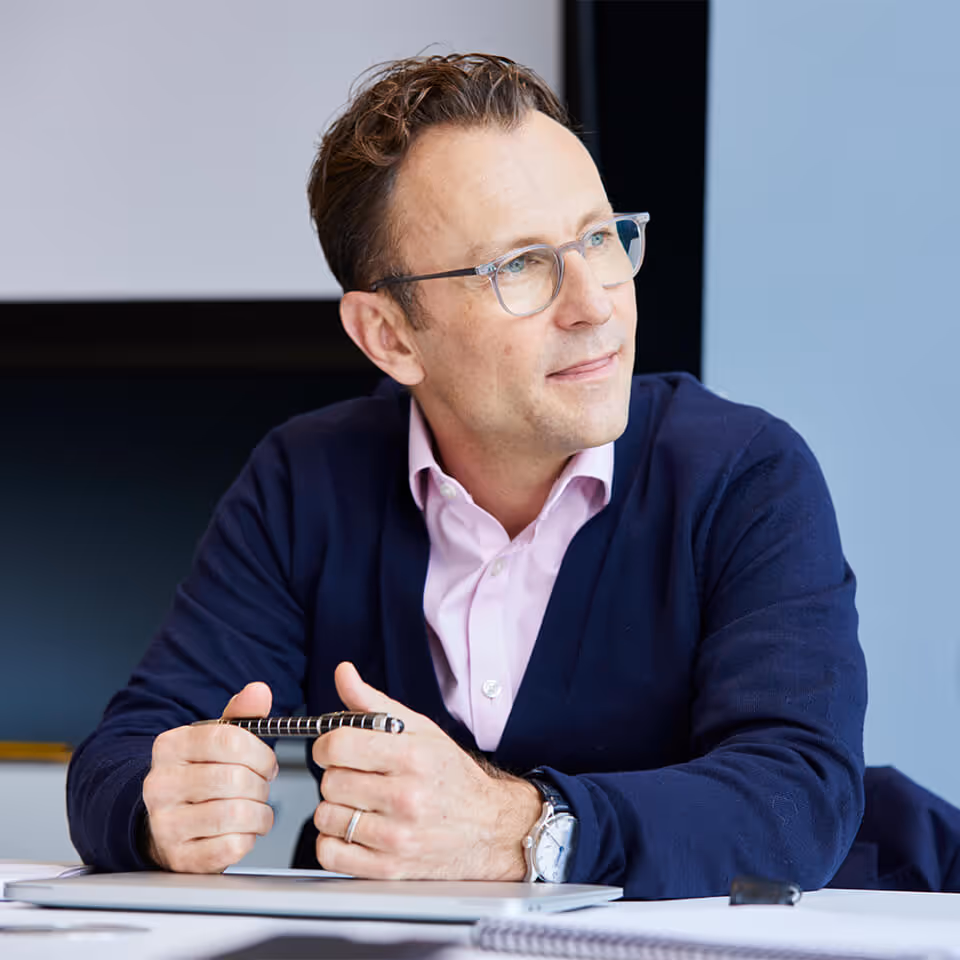 Man wearing glasses and a navy cardigan holding a pen, sitting at a table and looking thoughtfully to the side.