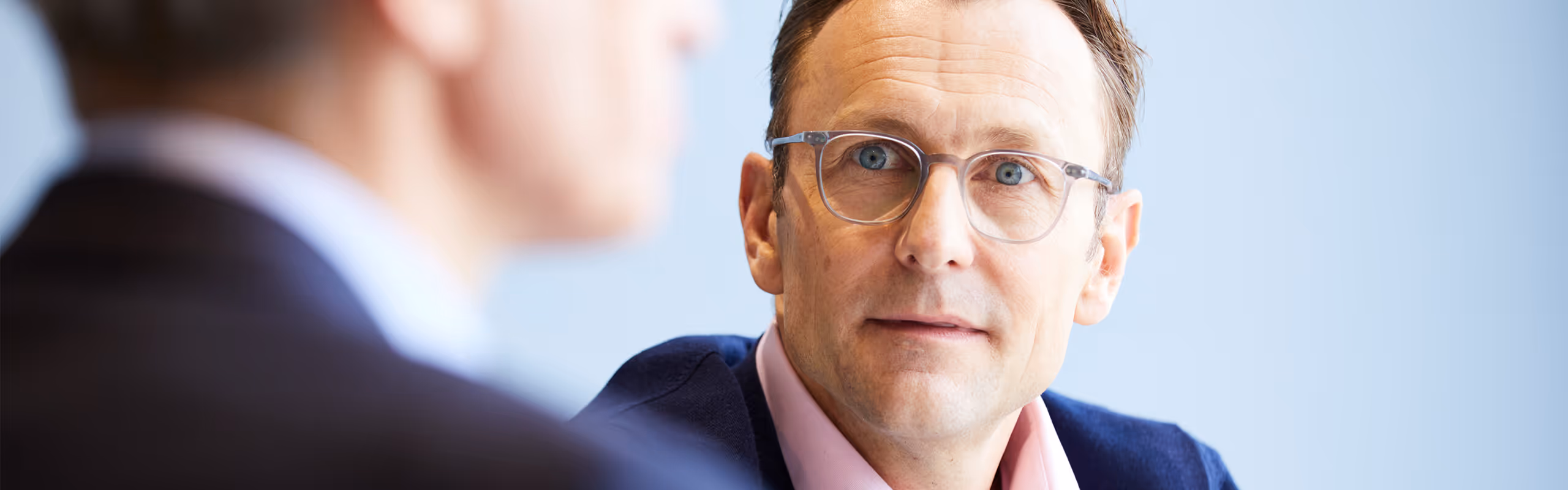 Close-up of a man with glasses and blue eyes attentively listening to another person in a suit in a professional setting.