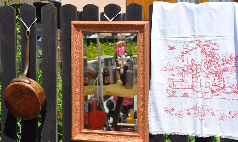 Un vieux poêlon en cuivre accroché à une clôture en bois sombre avec un miroir encadré et un tissu blanc brodé en rouge représentant un couple assis sur un banc.