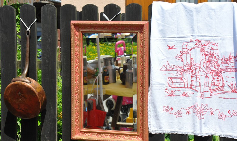 Un vieux poêlon en cuivre accroché à une clôture en bois sombre avec un miroir encadré et un tissu blanc brodé en rouge représentant un couple assis sur un banc.