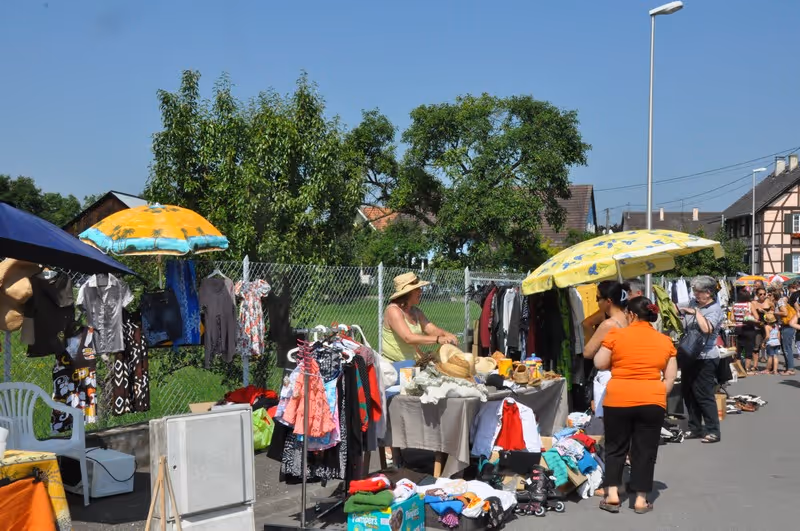 Marché en plein air avec stands de vêtements et accessoires, plusieurs personnes discutant et achetant sous un ciel clair.