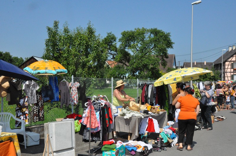 Marché en plein air avec stands de vêtements et accessoires, plusieurs personnes discutant et achetant sous un ciel clair.