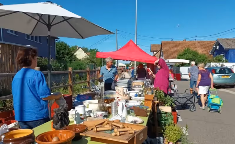 Marché de plein air avec des personnes regardant des objets divers sur une table sous un parasol blanc, avec une tente rouge en arrière-plan.