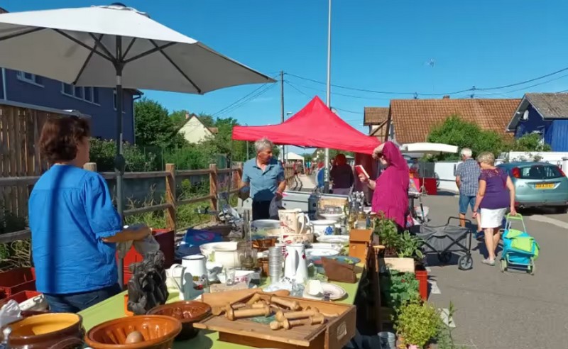 Marché de plein air avec des personnes regardant des objets divers sur une table sous un parasol blanc, avec une tente rouge en arrière-plan.