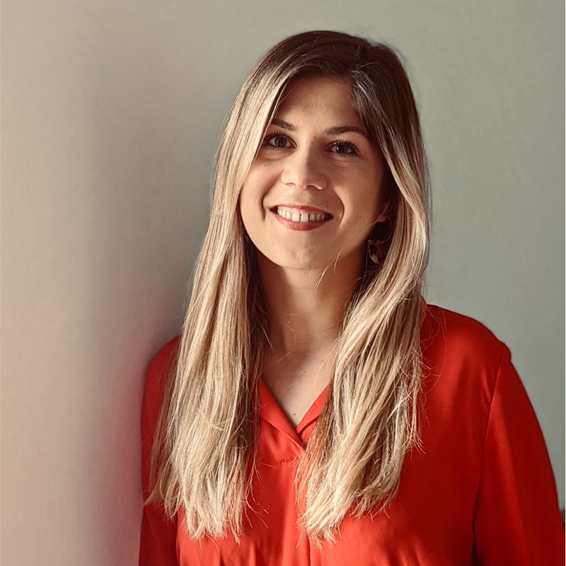 Smiling woman with long blonde hair wearing a red blouse standing against a light gray wall.