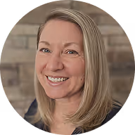 Smiling woman with straight blonde hair in front of a blurred brick wall.