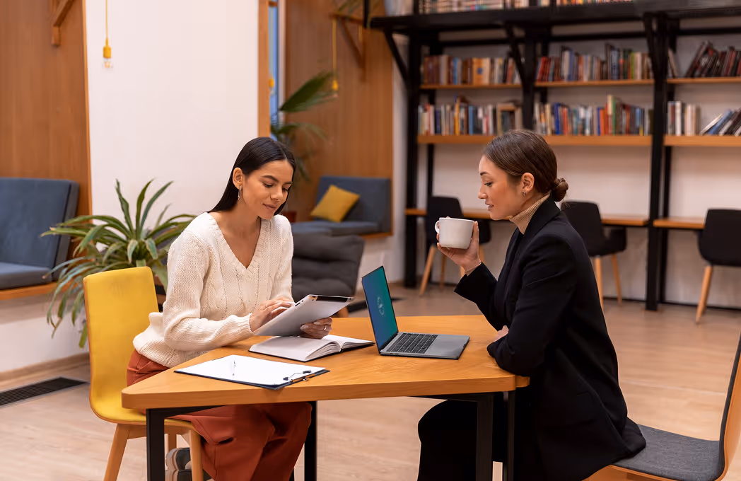 Two women seated at a wooden table in a modern library, one holding a tablet and the other drinking from a white mug, with bookshelves and plants in the background.