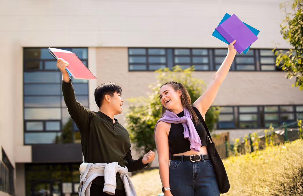 Two college students outdoors happily holding up colorful folders in front of a campus building.