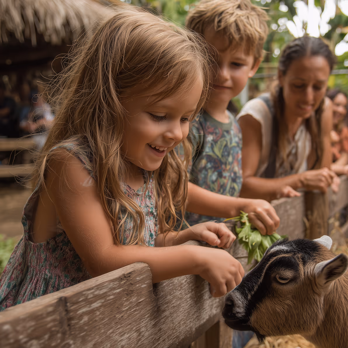 Smiling children and a woman feeding a goat over a wooden fence in an outdoor setting.