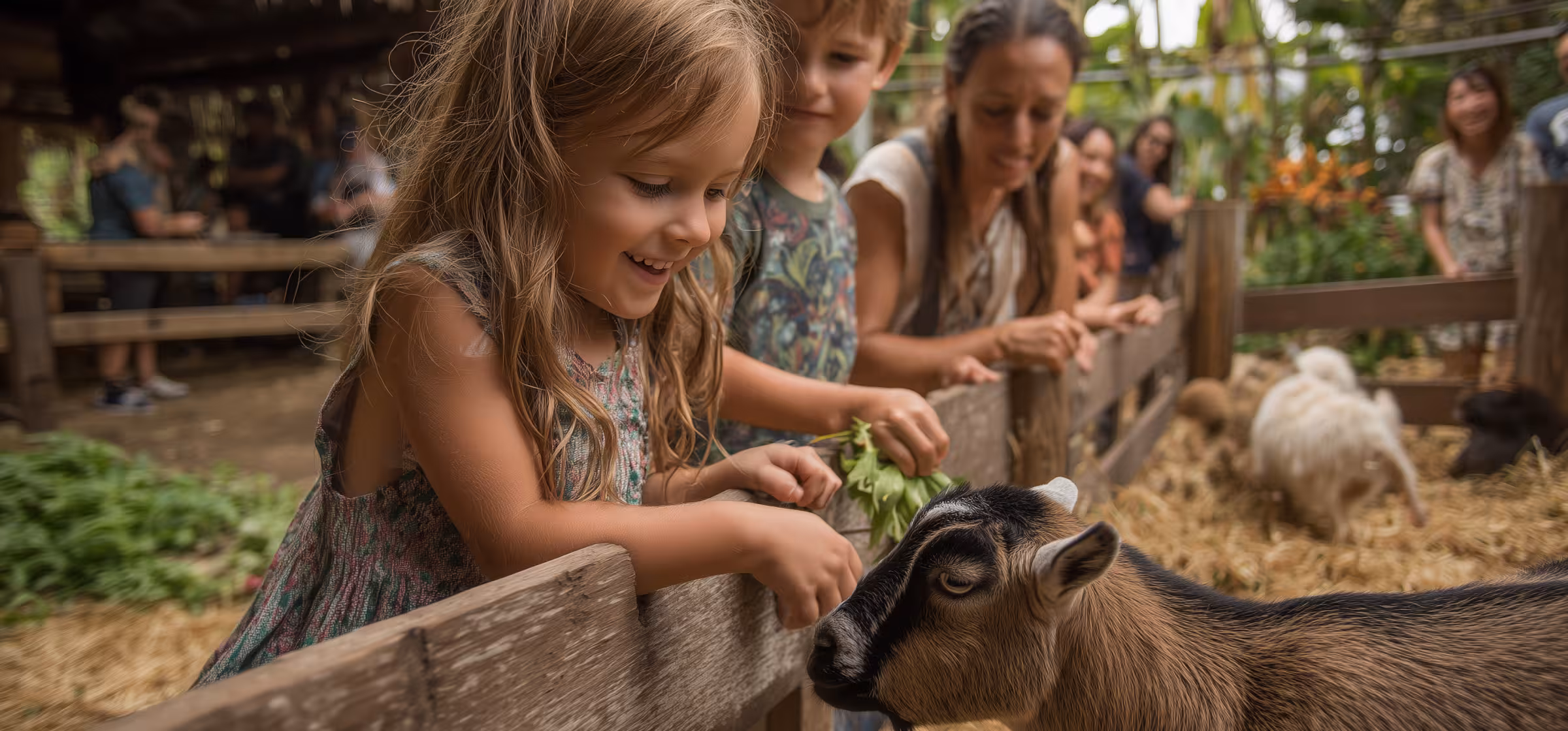 Young girl smiling while feeding a brown and black goat over a wooden fence at a petting zoo.